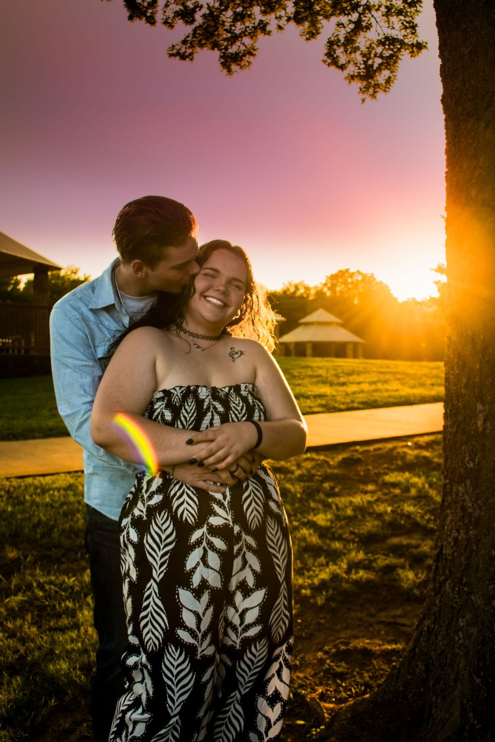 A romantic couple embraces outdoors during golden hour sunset with lens flare and trees in the background.