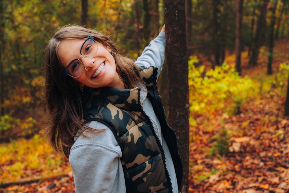 A person in a gray sweater poses playfully against autumn trees with vibrant orange and yellow foliage in the background.