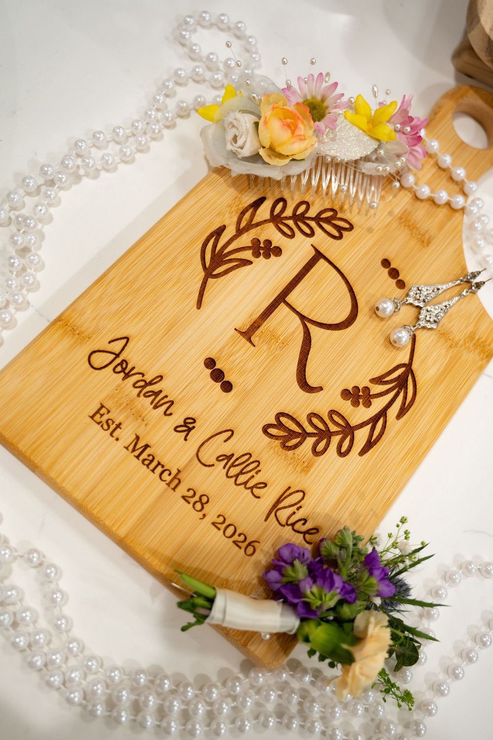 Personalized engraved bamboo cutting board for Jordan & Callie Rice, Est. March 28, 2026, surrounded by pearls and flowers.