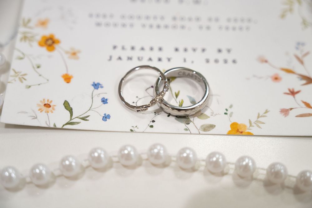 Two silver wedding bands resting on a floral RSVP card beside a pearl necklace on a white surface.