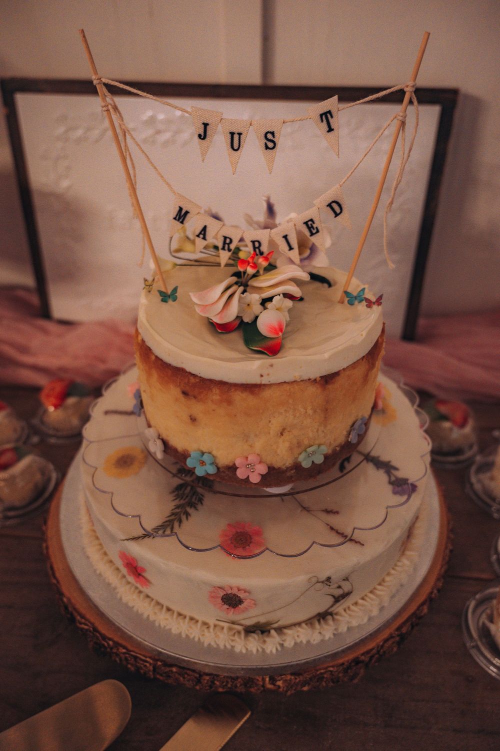 Three-tier wedding cake with floral decorations and a rustic wood base, topped with a charming Just Married banner topper.