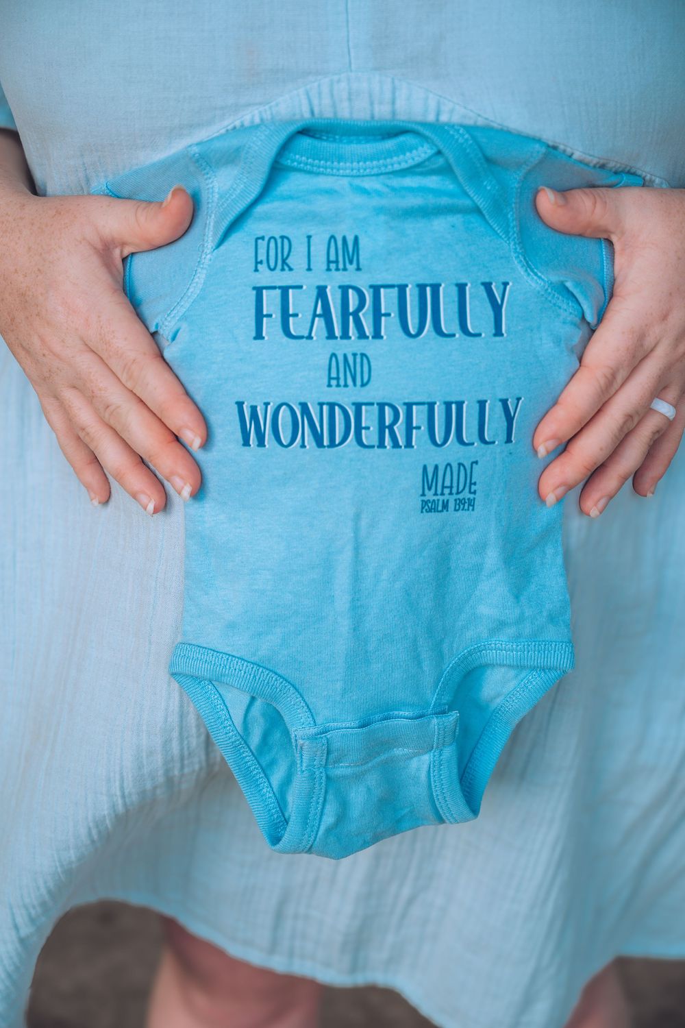 A pregnant woman holds a blue baby onesie reading For I Am Fearfully and Wonderfully Made Psalm 139.