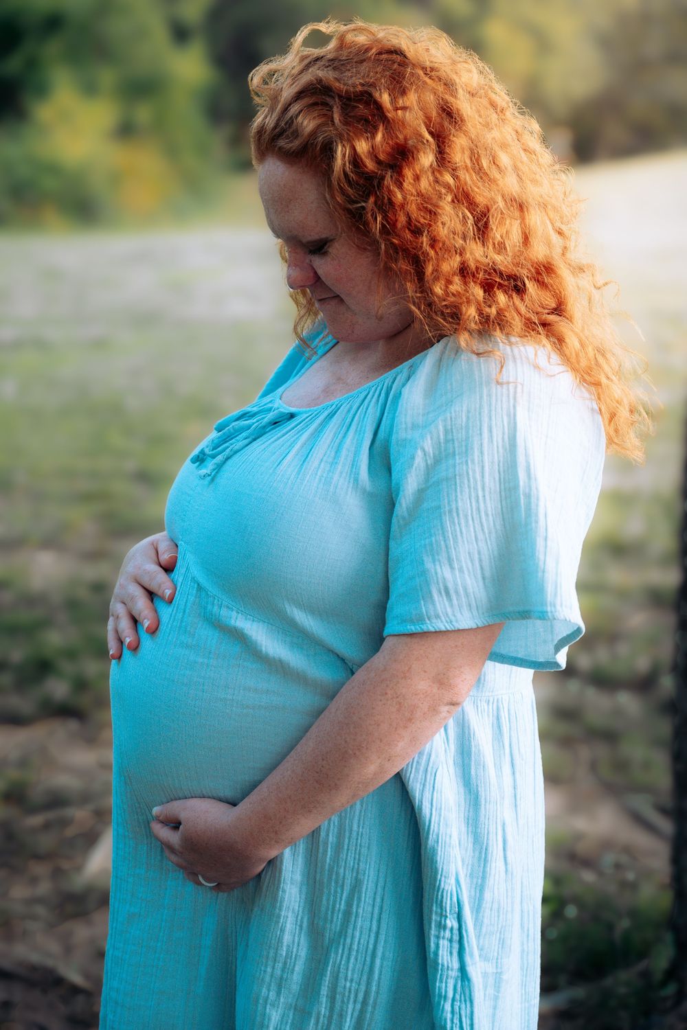 Pregnant redhead woman in teal dress cradling baby bump outdoors in natural setting.