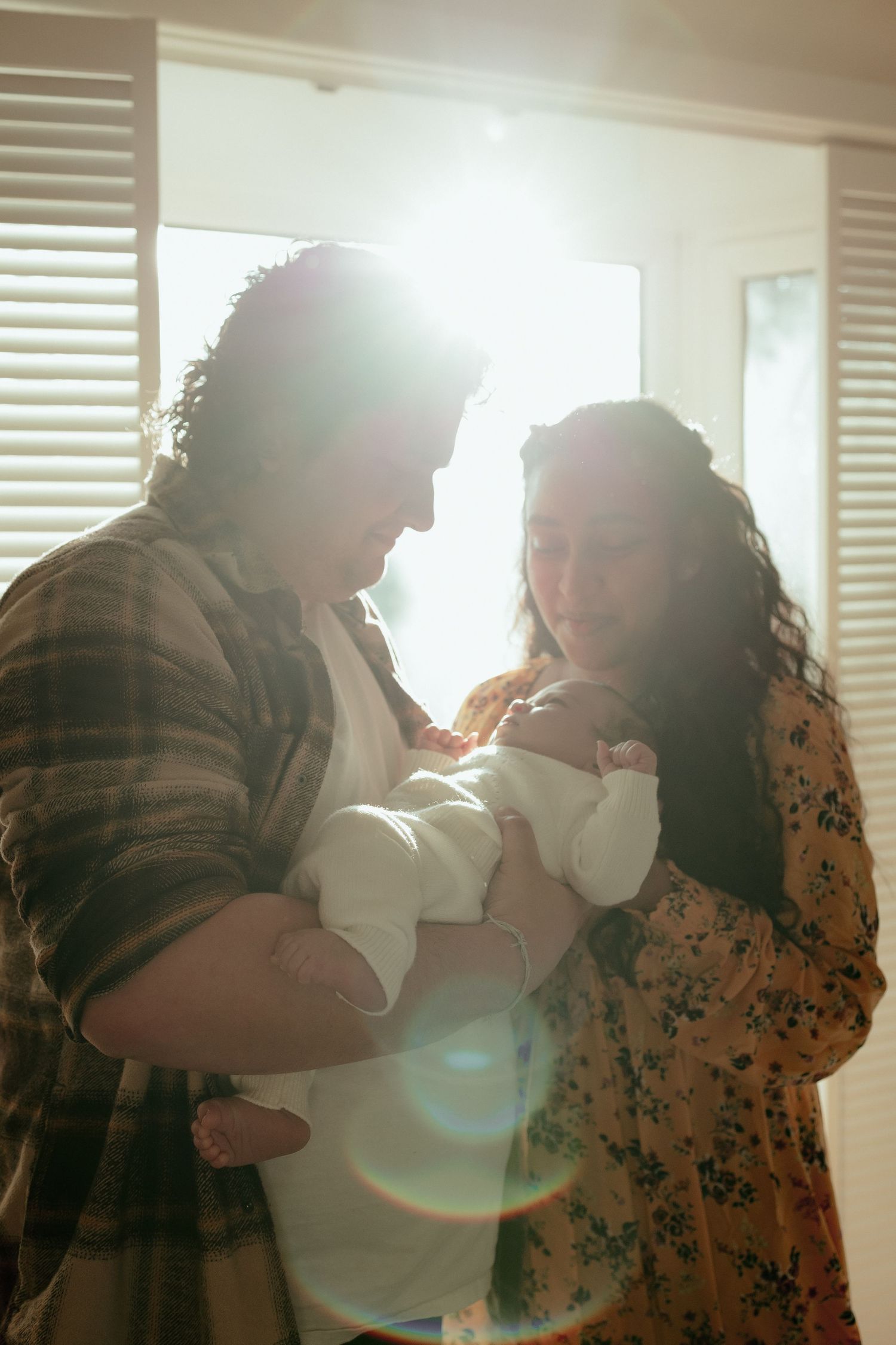 Parents hold their newborn baby in a sunlit room with window blinds casting a warm glow.