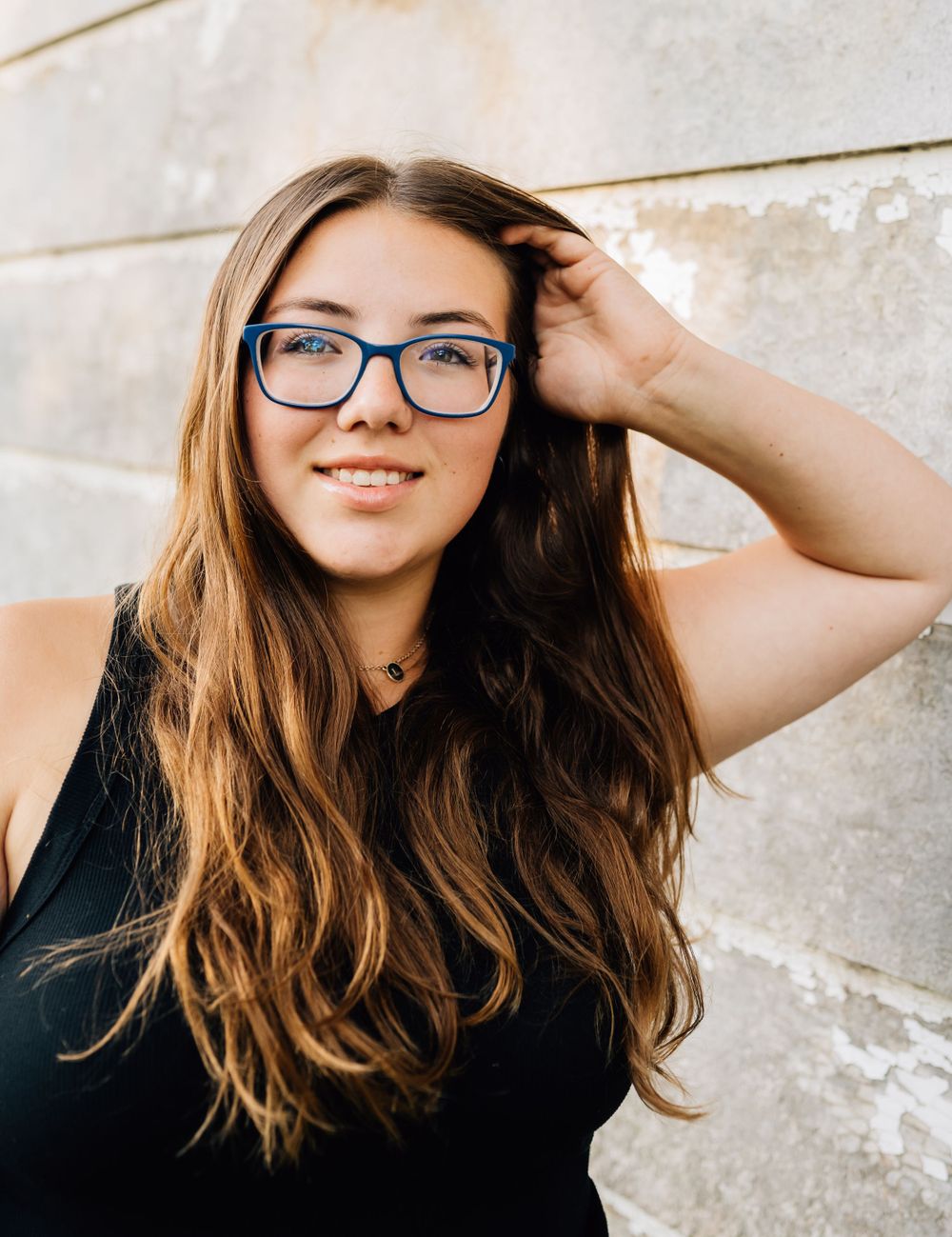 Portrait of someone in glasses and black top posing against light wall with long wavy hair.