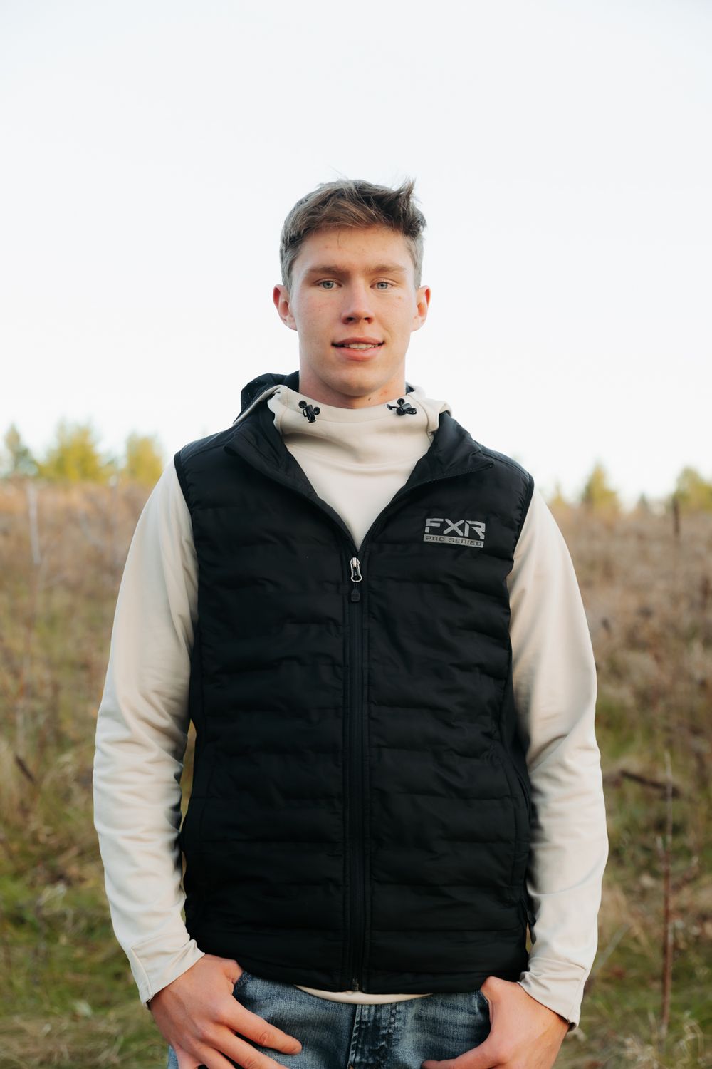 A casual portrait in a field wearing a black vest and white turtleneck sweater.