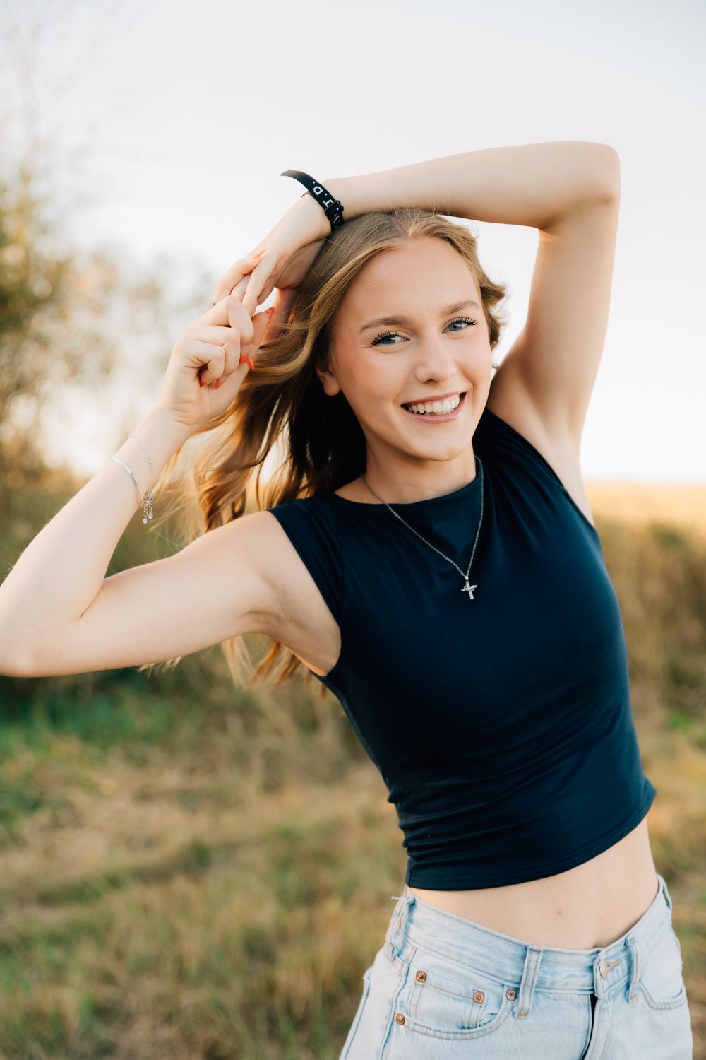Young woman in black crop top and jeans poses playfully outdoors during golden hour sunset.