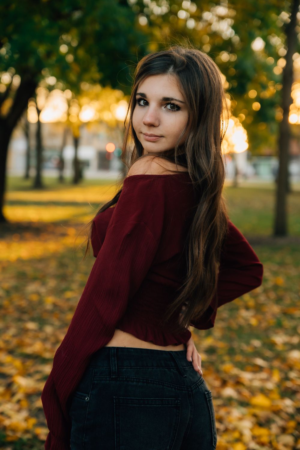 Portrait of someone in a burgundy crop sweater posing in a park with golden autumn leaves at sunset.