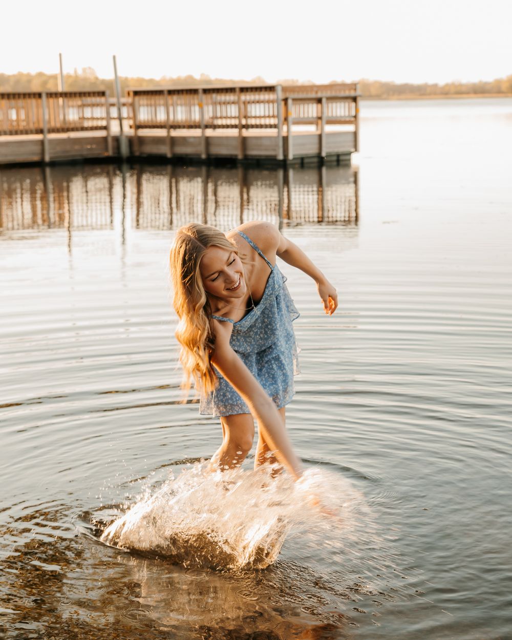 Woman in blue dress splashing water at lake dock during golden hour.