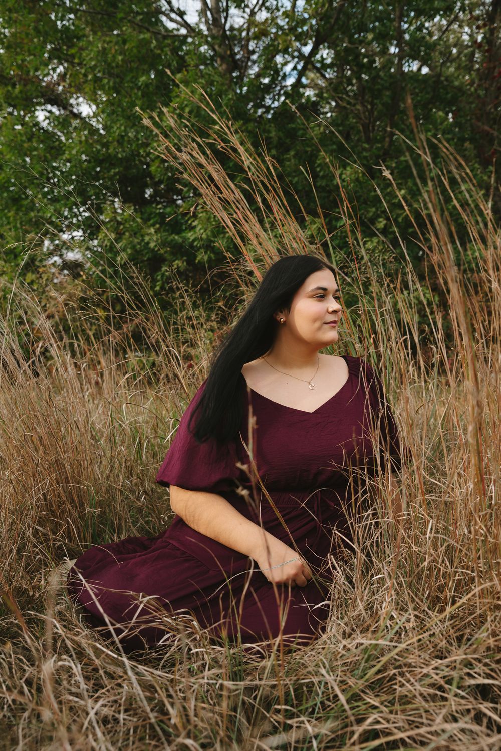 Woman in burgundy dress sitting among tall grass in natural outdoor setting.