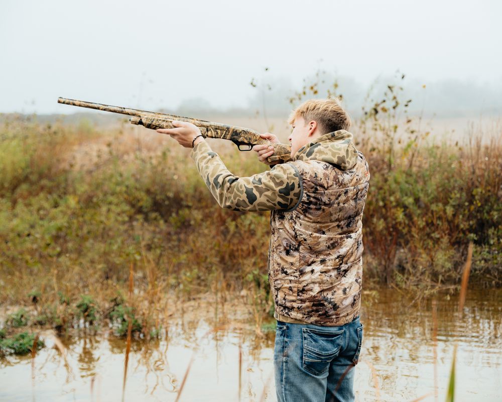 Hunter in camouflage gear aiming shotgun while duck hunting near water.