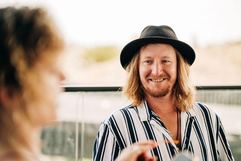 Smiling man with long hair wearing a black hat and striped shirt outdoors.