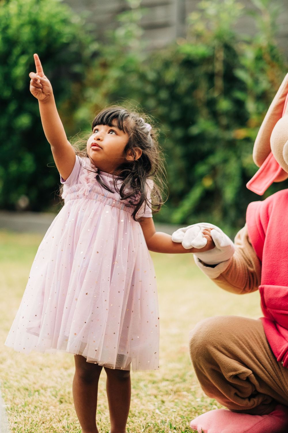 A young girl in a pink tutu dress points upward while holding hands with an adult outdoors.