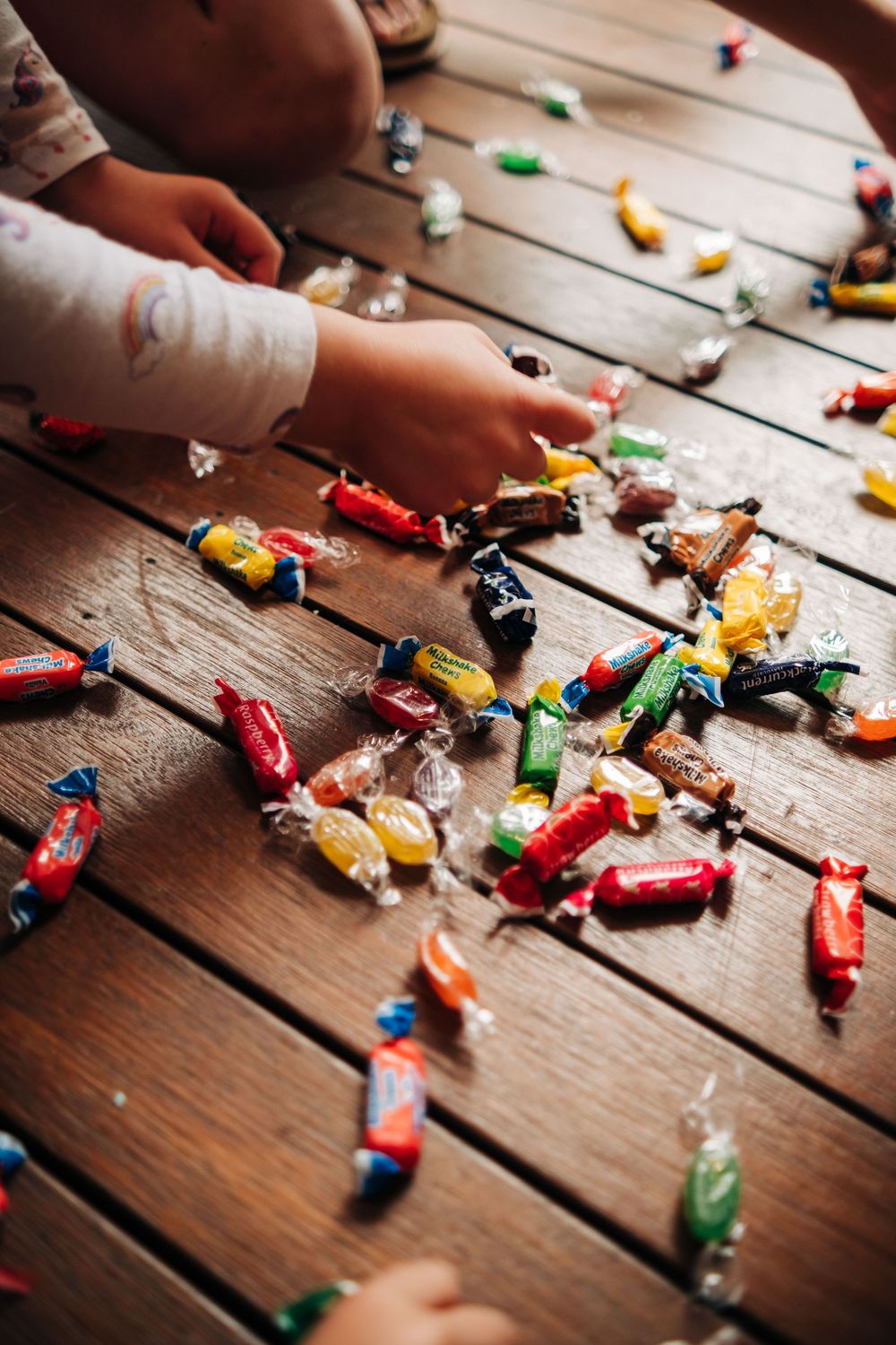 Children's hands reaching for colorful wrapped candies scattered on a wooden floor.