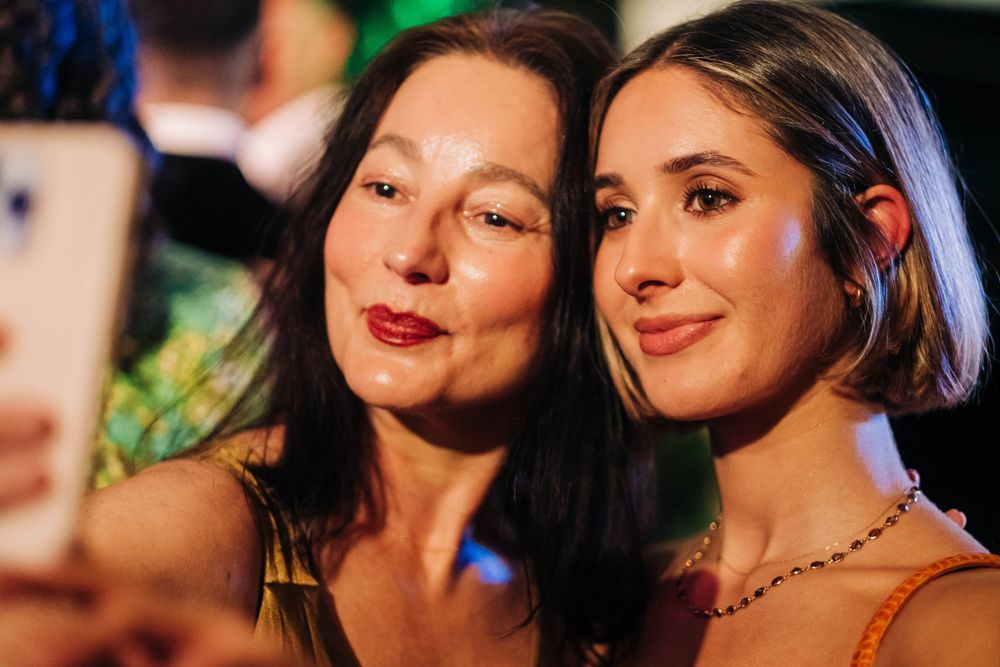Two women pose for a selfie at a glamorous evening event, smiling warmly under colorful ambient lighting.