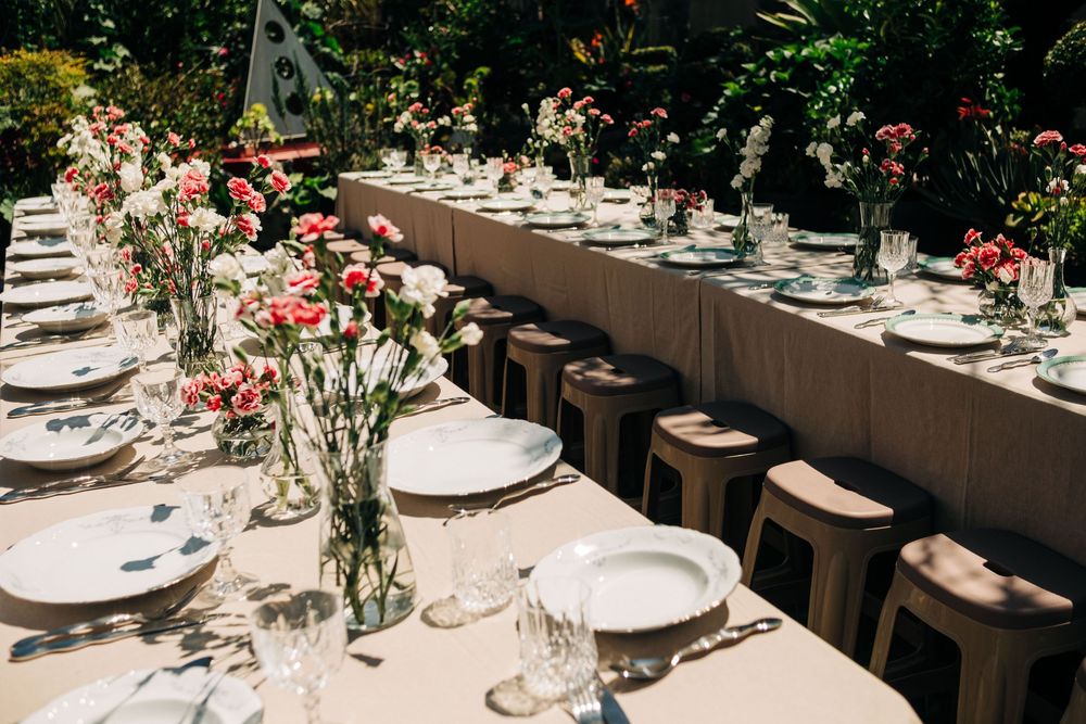 Elegant outdoor long dining table set with pink floral arrangements, white plates, and glassware for a garden wedding reception.