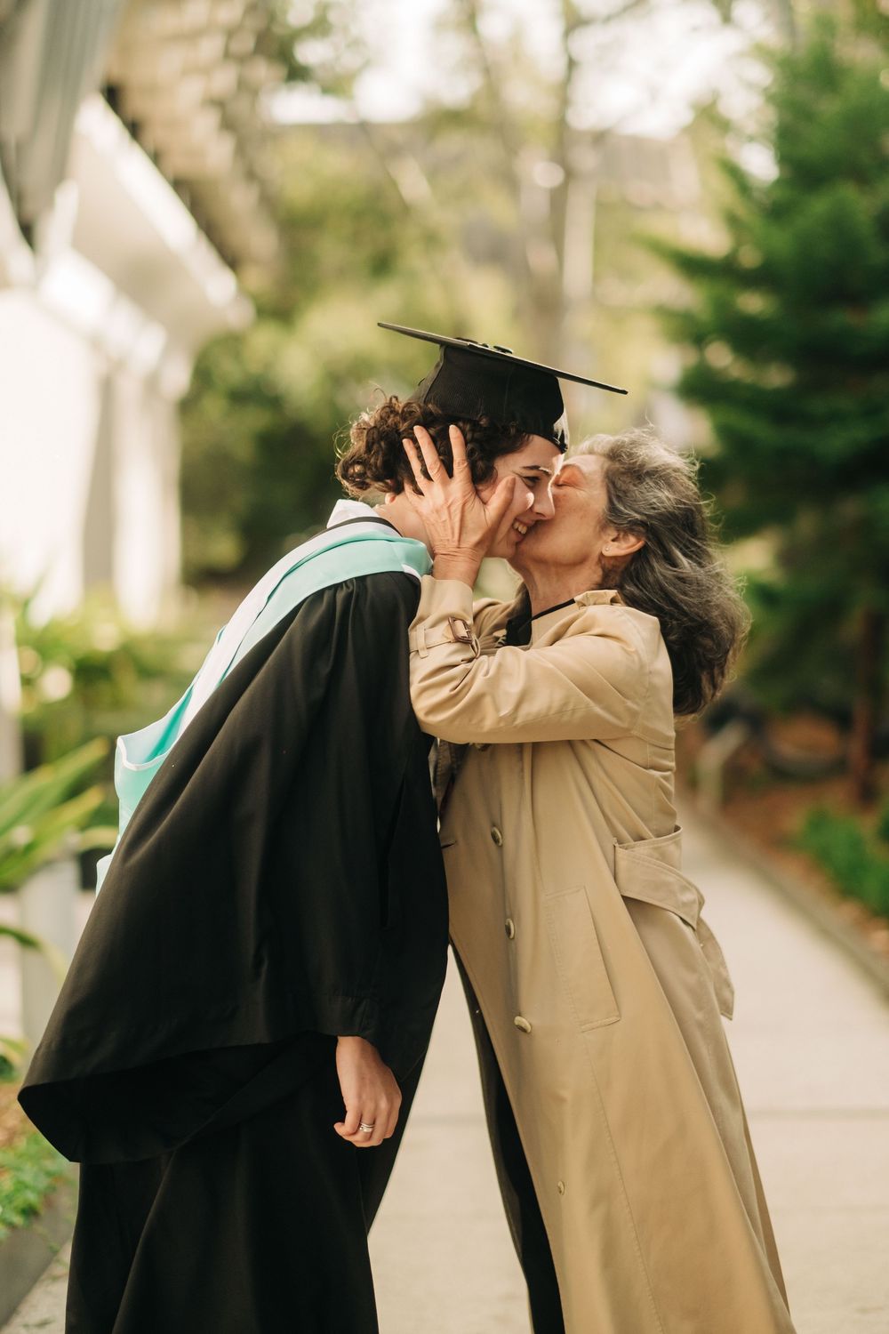 A proud mother kisses her child in graduation cap and gown outdoors on a campus pathway.