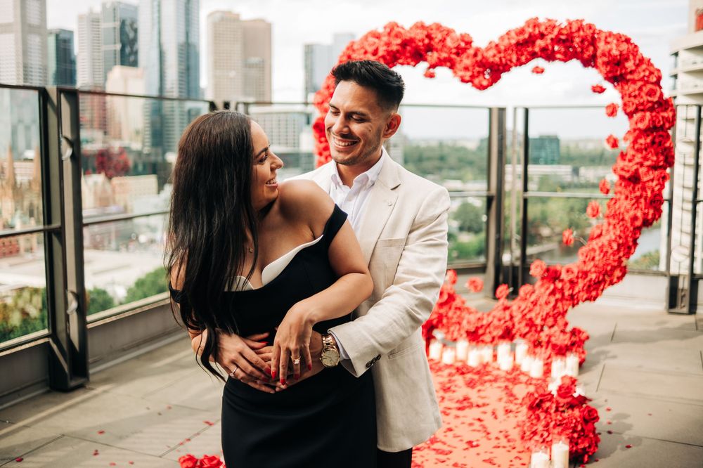 Couple embracing in front of a red rose heart arch on a rooftop, celebrating a romantic proposal.