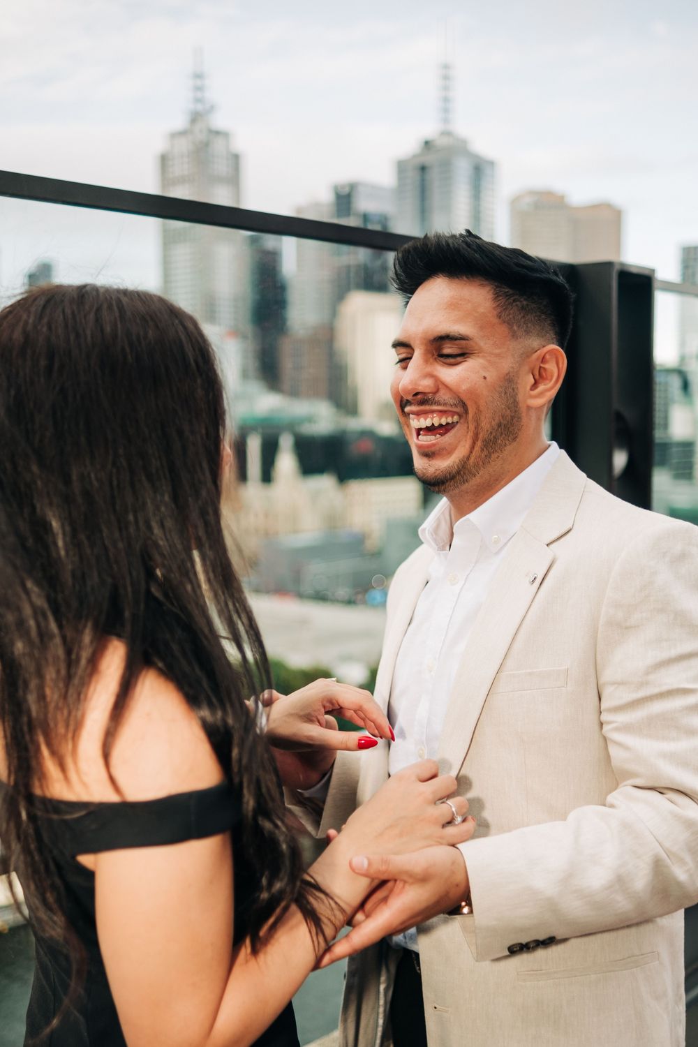 Couple sharing a joyful moment on a rooftop terrace with city skyline backdrop, man in white suit laughing.