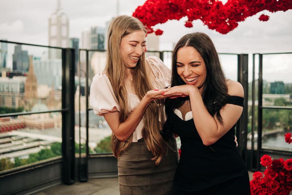 Two women smiling on a rooftop terrace with red floral decor and city skyline, sharing a joyful moment together.