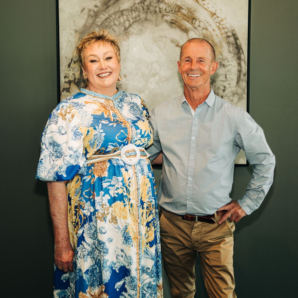Two smiling adults pose together in front of abstract wall art, woman in floral blue dress, man in light blue shirt.