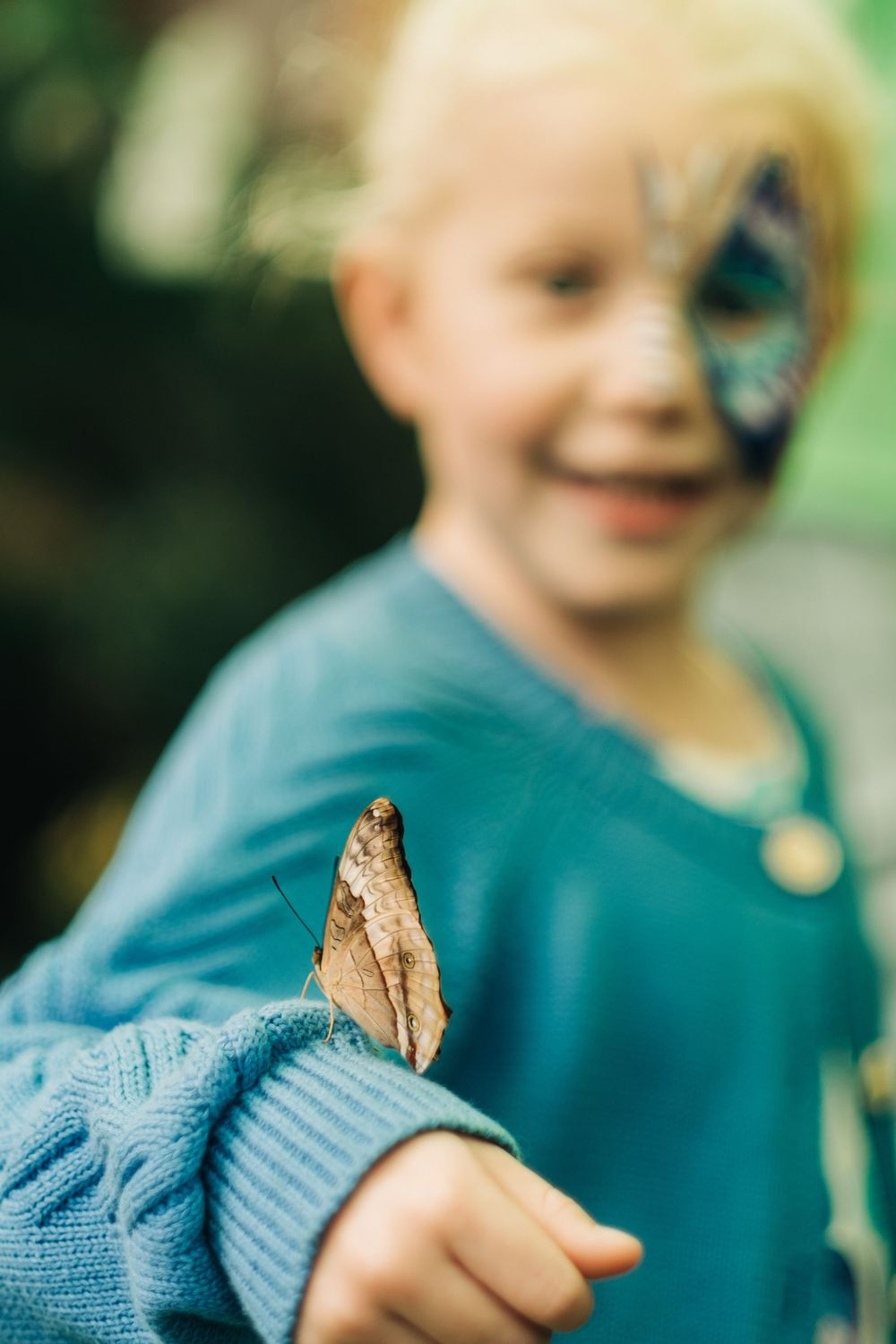 A smiling blond child with butterfly face paint holds a brown butterfly on their outstretched arm outdoors.