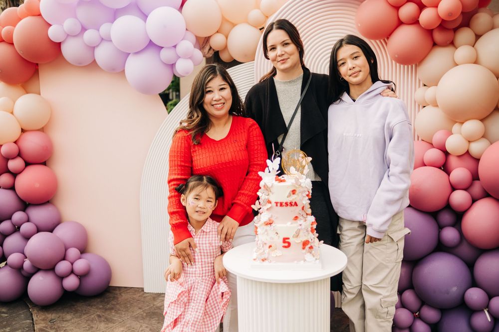 Family posing together at a birthday party with colorful balloon arch and a decorated 5th birthday cake.