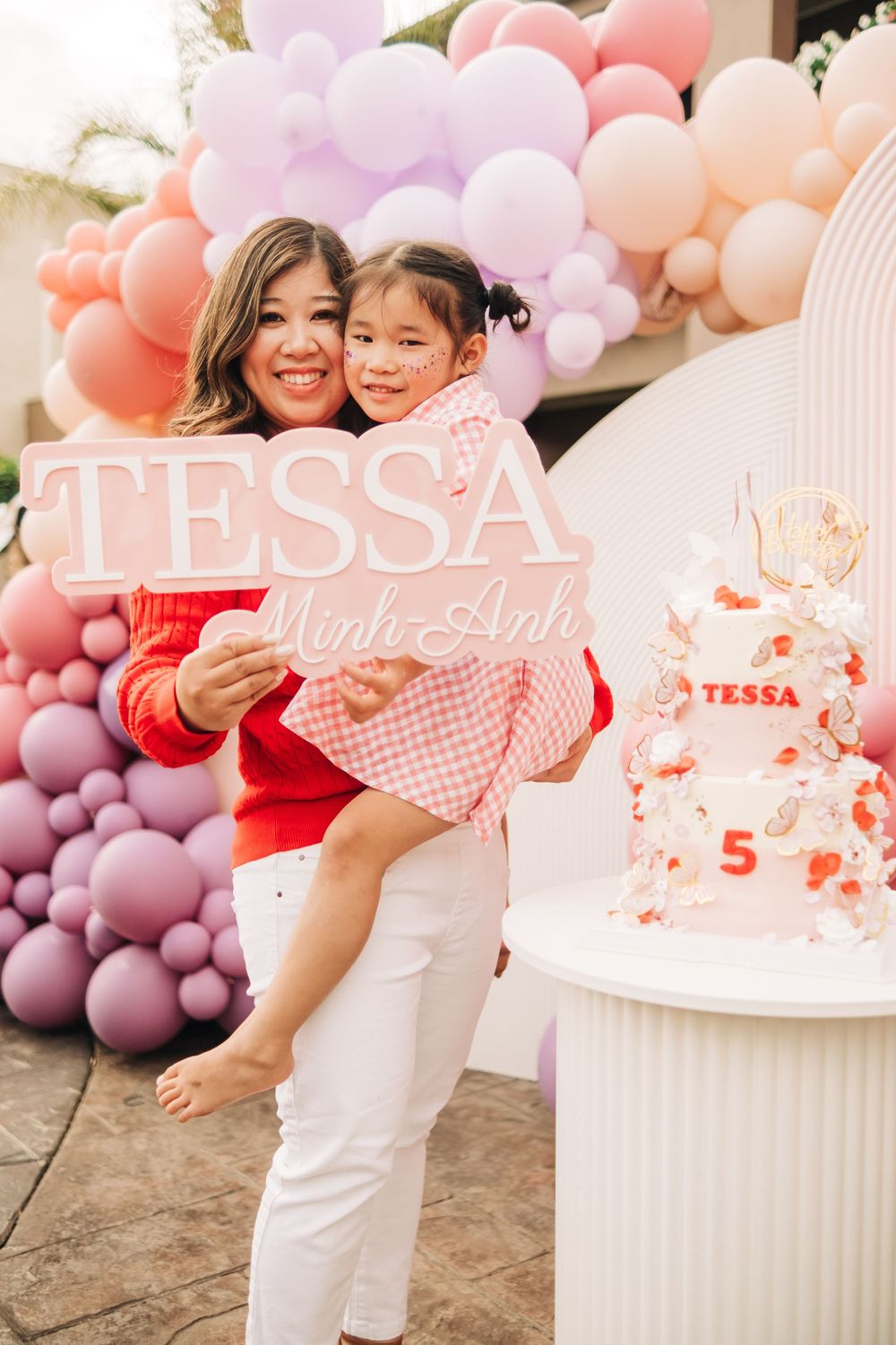 Mom holds daughter Tessa at her 5th birthday party with pink balloon arch and decorated cake.