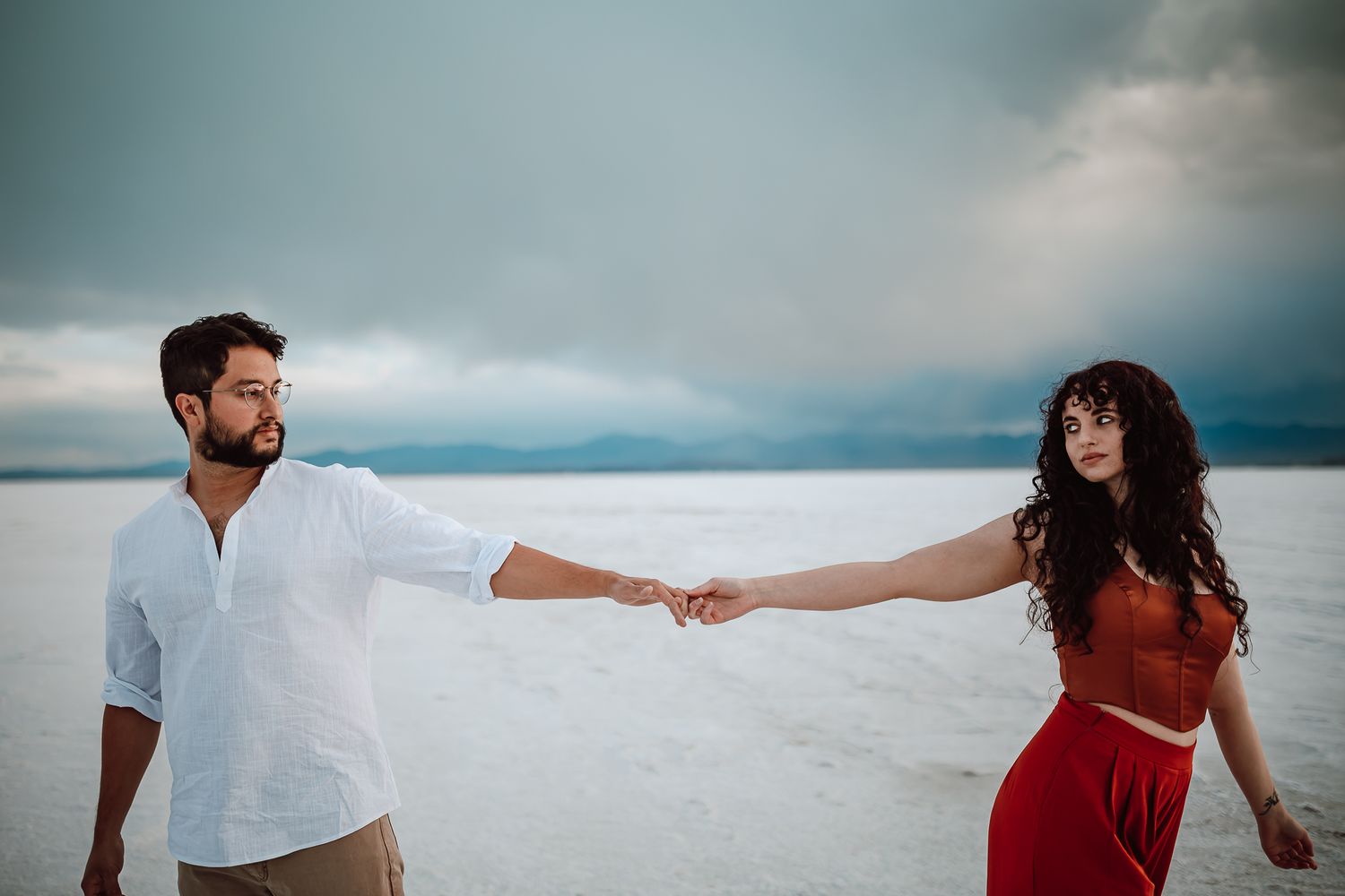 Couple in white shirt and red dress hold hands while standing on a white sand beach under dramatic cloudy skies.