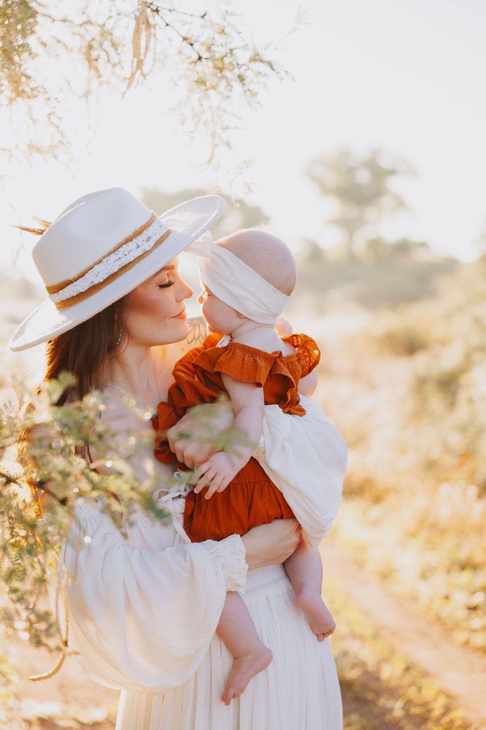 Mother and child in white dresses and hats embracing in sunlit desert landscape.