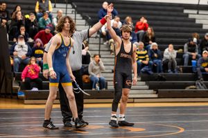 Wrestling match in progress on orange mat with spectators watching from bleachers in background.