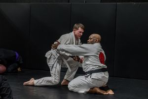 Two martial artists practicing Brazilian Jiu-Jitsu grappling techniques on a black mat.
