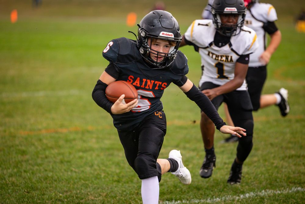 Youth football players in black uniforms running plays during a game on grass field.