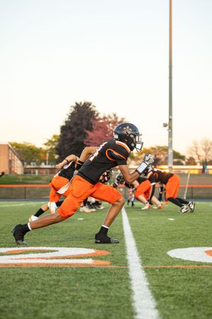Football player in orange uniform sprints forward during practice on a turf field at sunset.