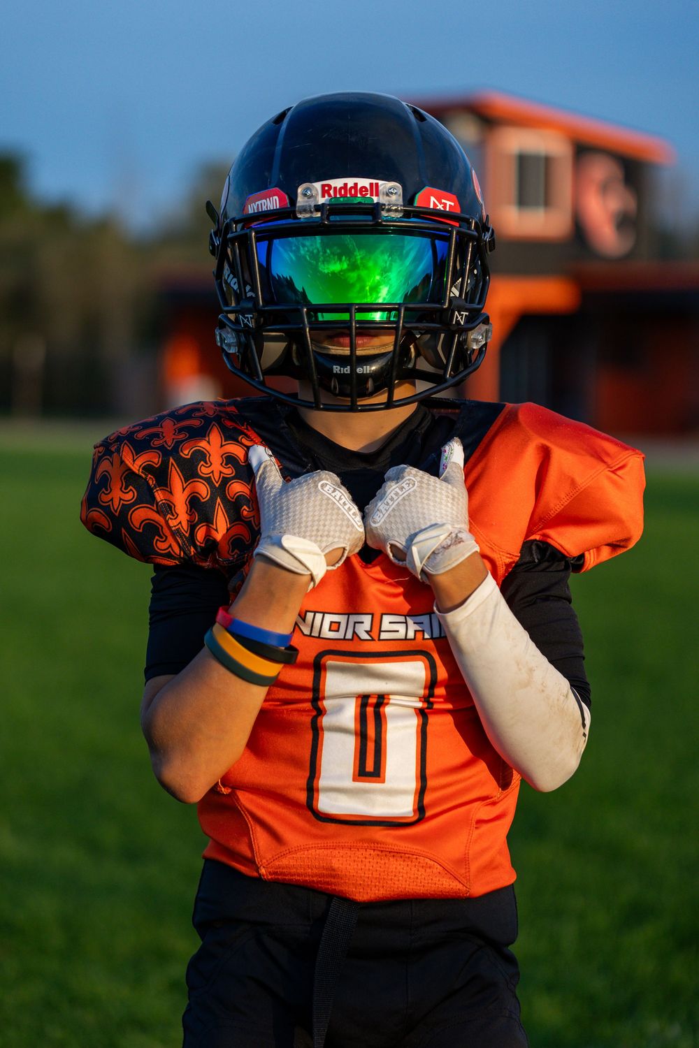 Youth football player in orange jersey poses with ball wearing helmet with green visor.