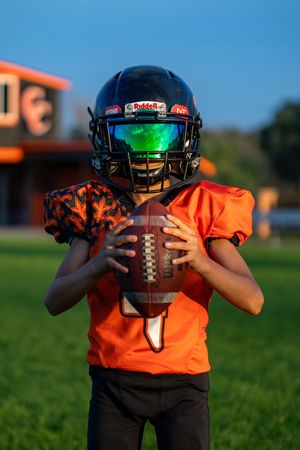 Young athlete in orange uniform holds football while wearing helmet with green visor.