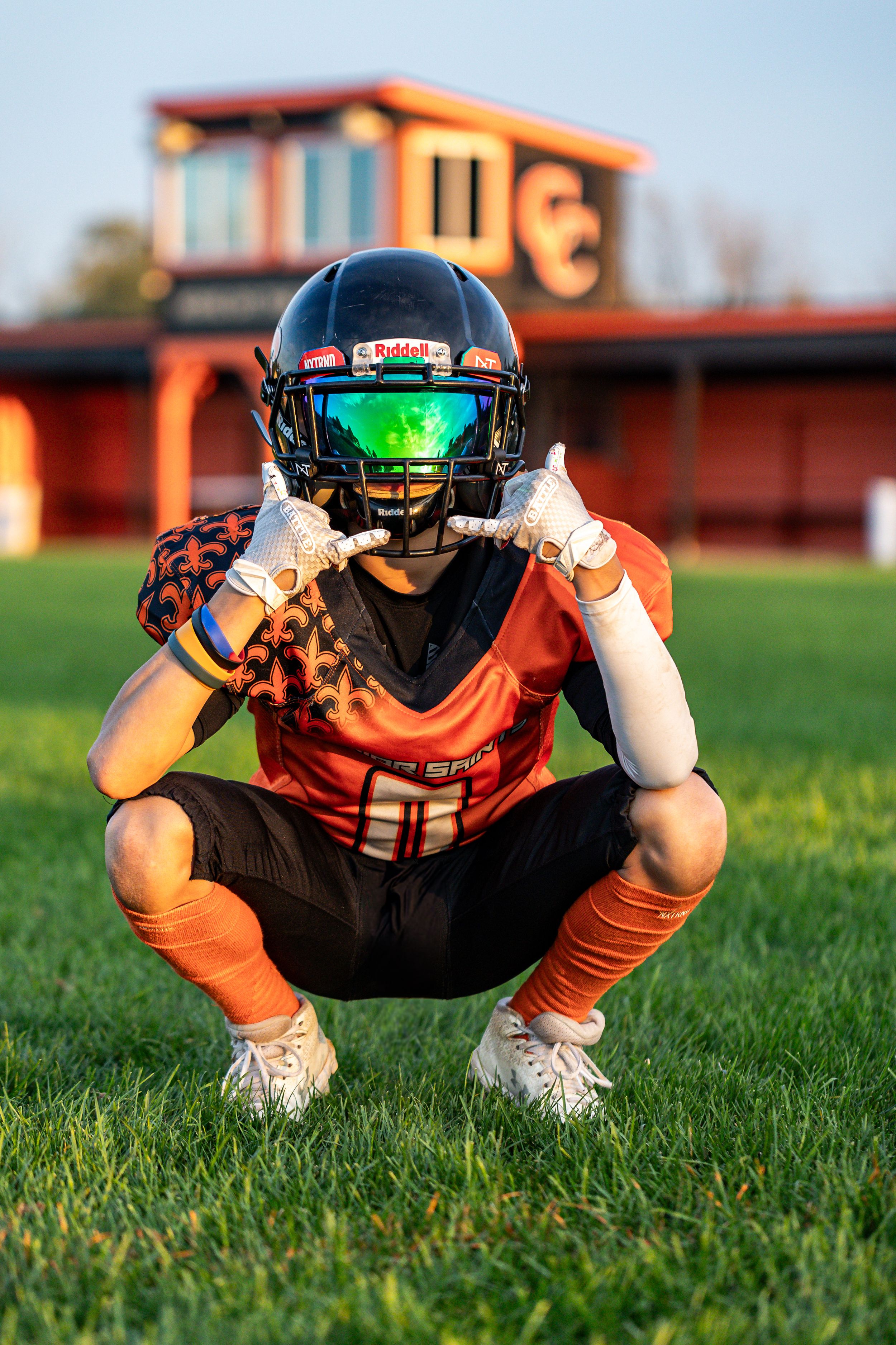 Football player crouches low on grass field wearing orange uniform and reflective visor helmet.