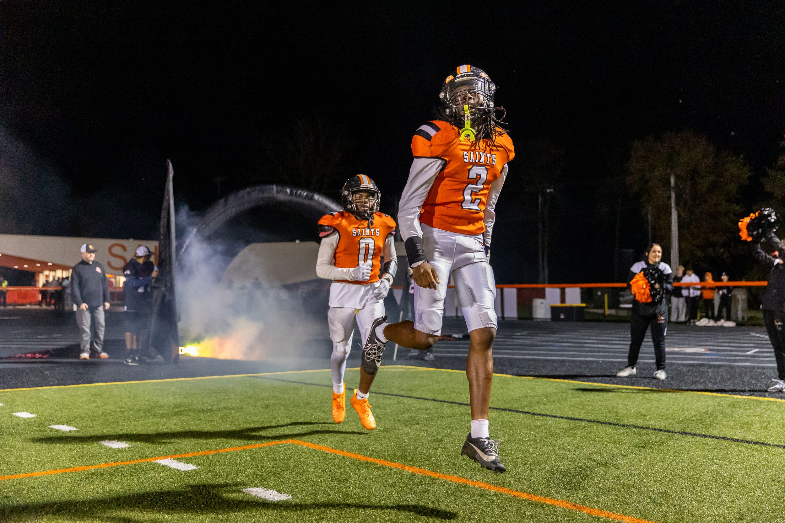 Two football players celebrate on the field at night with smoke effects in the background.