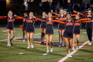 Cheerleaders perform synchronized routine with orange pom poms on football field.
