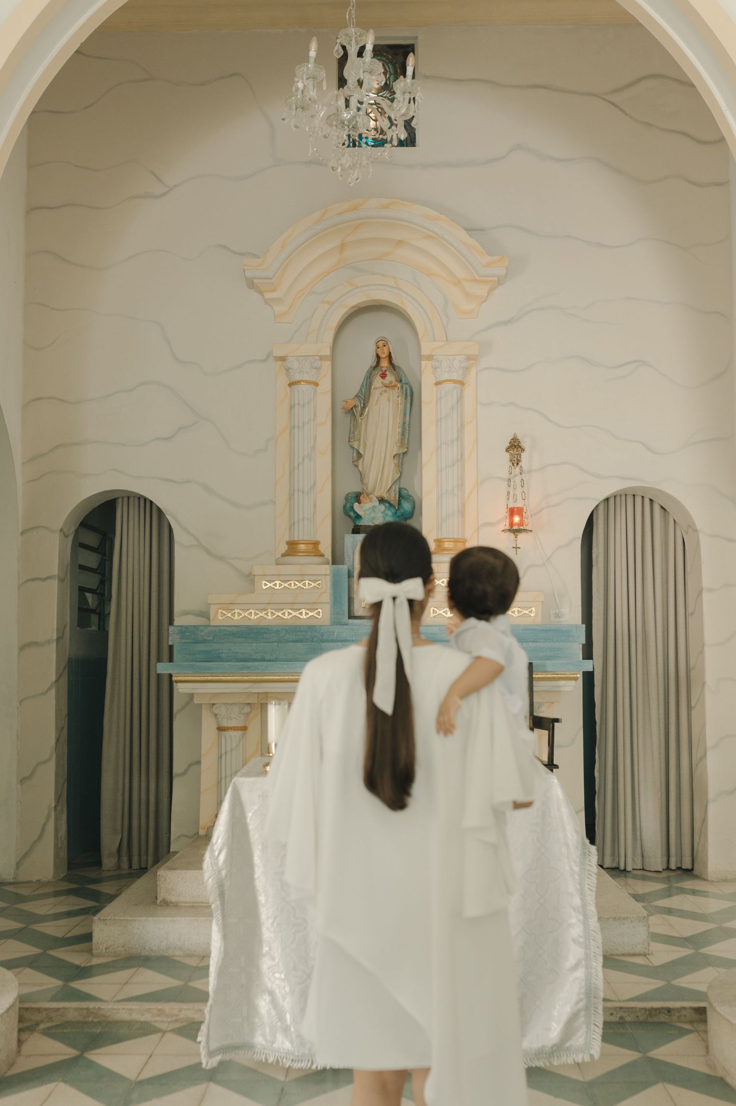 Couple in white robes standing before an ornate church altar with a statue of the Virgin Mary.