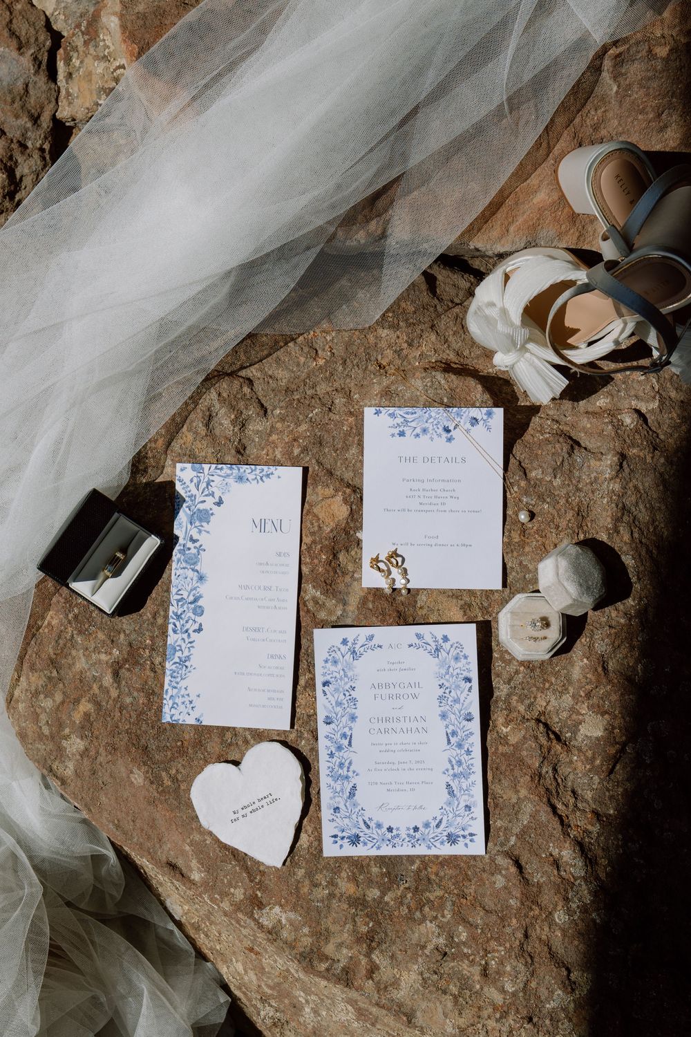 Elegant wedding invitation suite with delicate blue floral designs laid out on textured stone with flowing white tulle fabric.