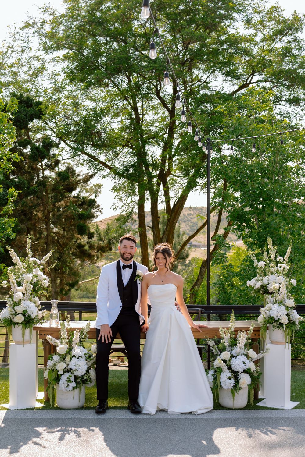 A romantic wedding couple poses between elegant white floral arrangements on pedestals at an outdoor venue.