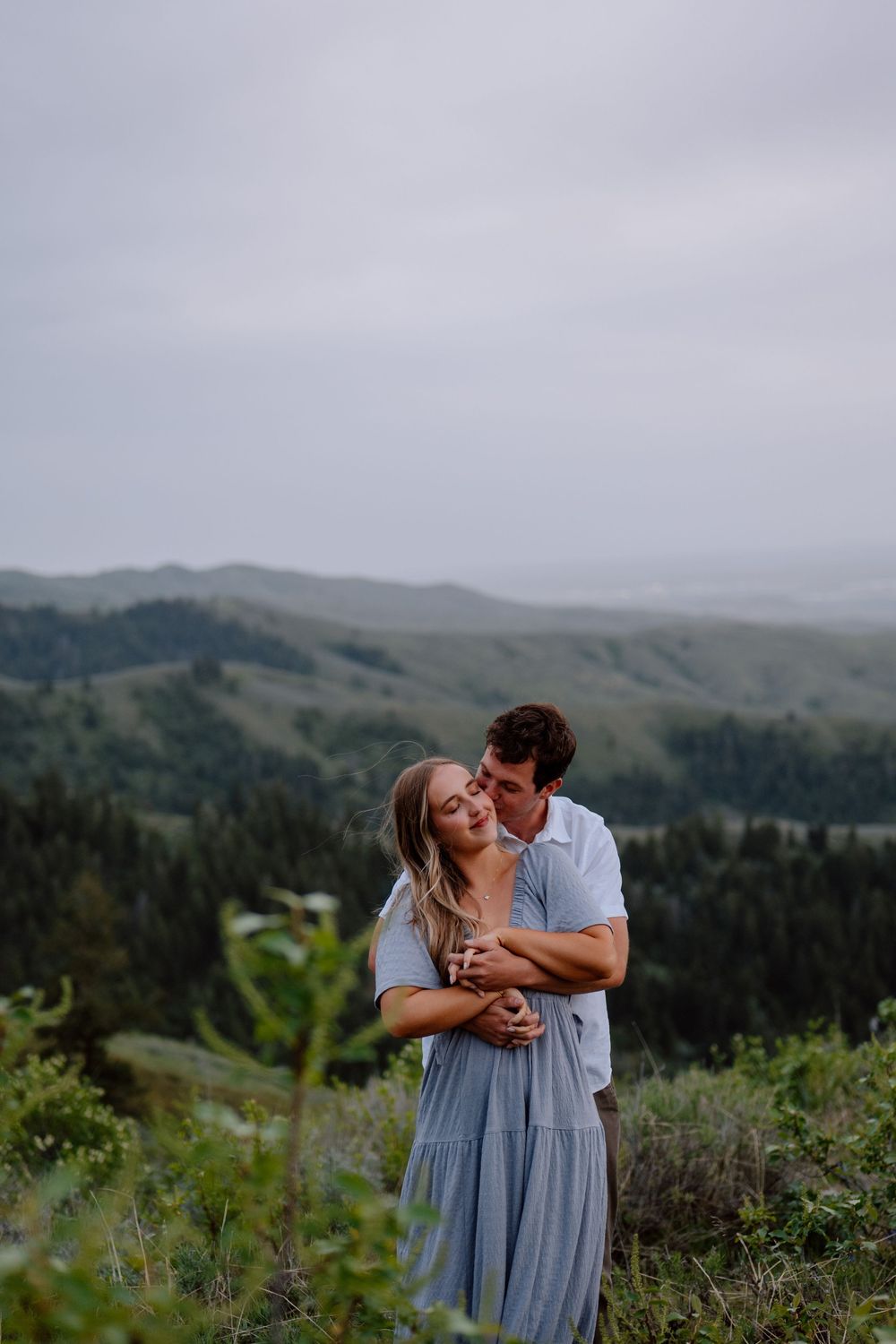 A couple embraces on a hilltop overlooking rolling mountains and forests on an overcast day.