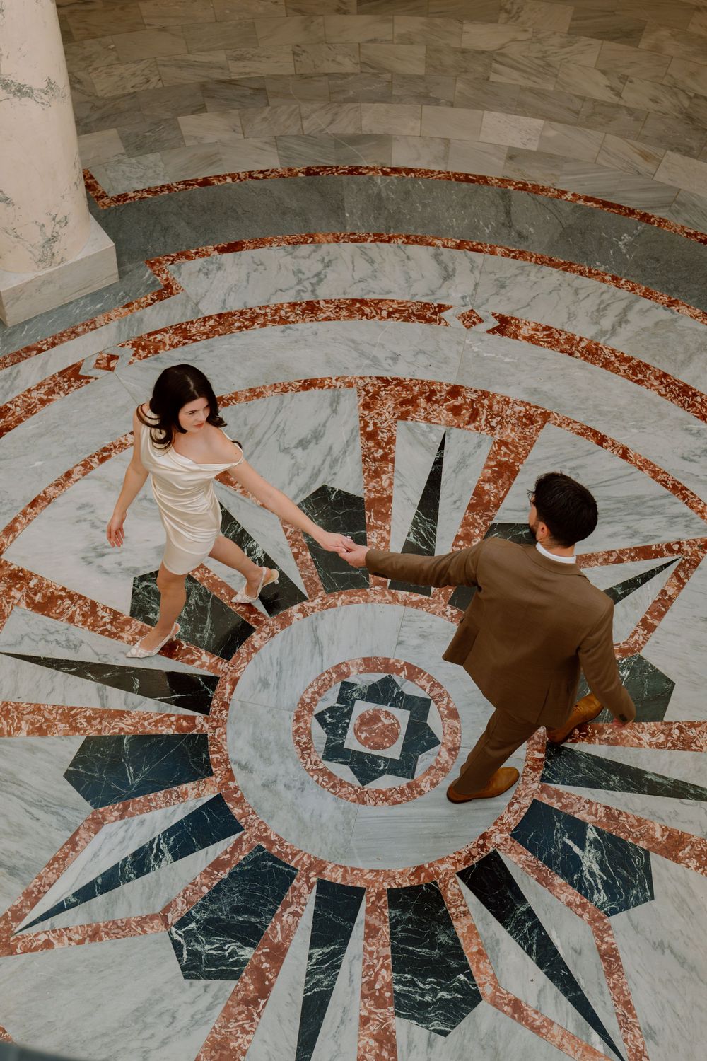 Couple holding hands while standing on ornate marble floor with geometric sunburst pattern in warm tones.