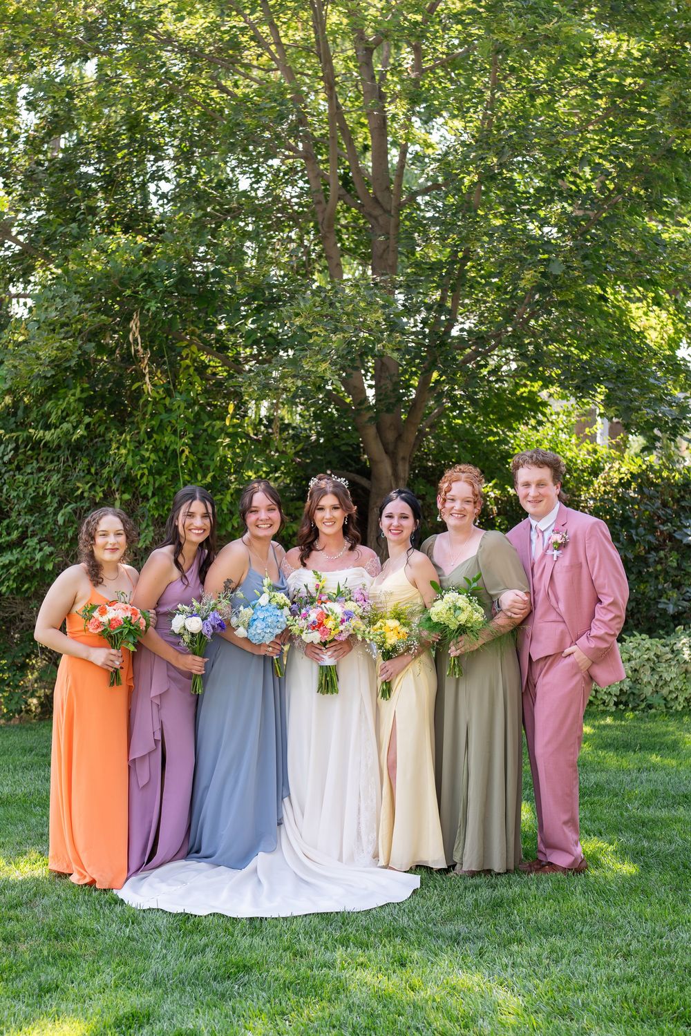Wedding party poses together on green lawn with bridesmaids in pastel dresses holding colorful bouquets.