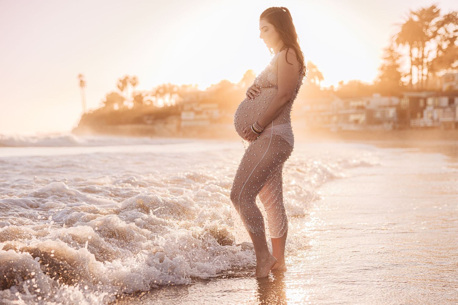 Silhouetted pregnant figure stands at the ocean's edge during golden sunset as waves lap at her feet.
