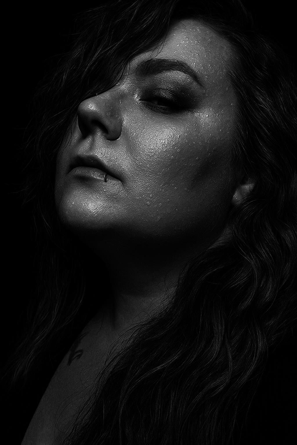 Black and white dramatic portrait of a woman with water droplets on her face, gazing upward against a dark background.