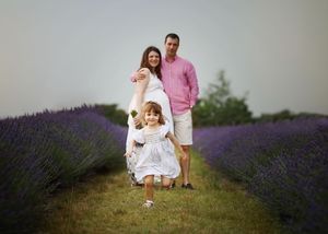 Family of three posing in a lavender field, parents and young daughter in white dress smiling.