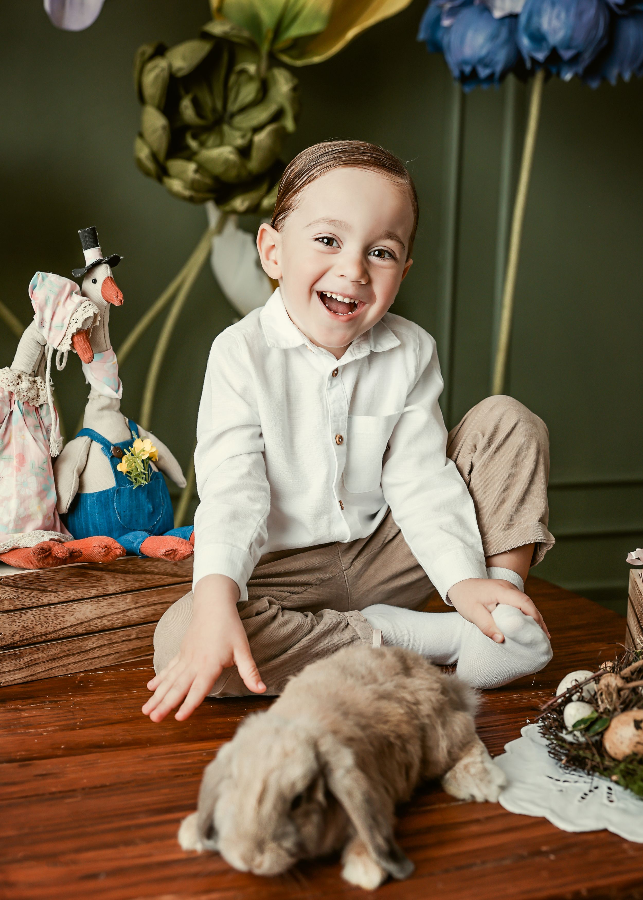 Laughing boy in white shirt sits on wooden table beside a rabbit and Easter decorations.