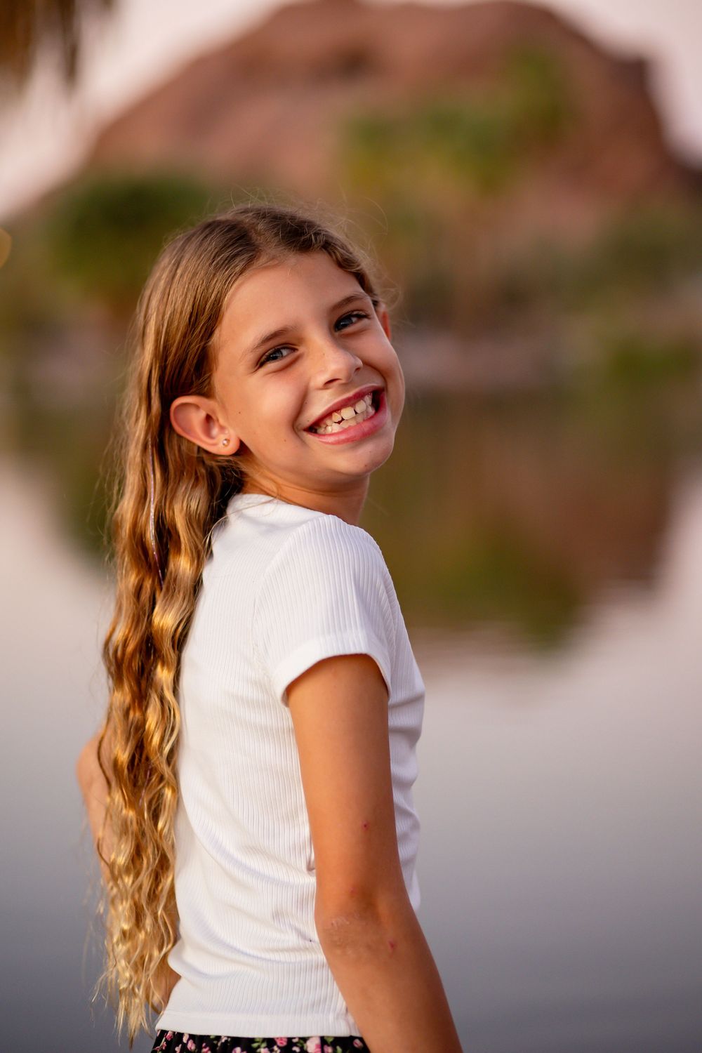 Young blonde girl with long braided hair smiling outdoors near a reflective lake at golden hour.